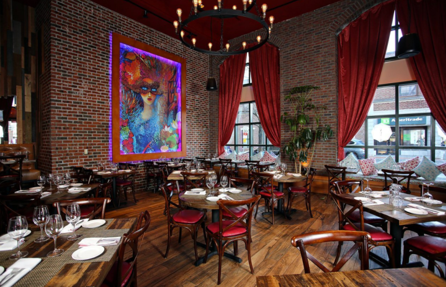 Restaurant interior with brick walls, red accents, and large artwork. Tables set for dining.