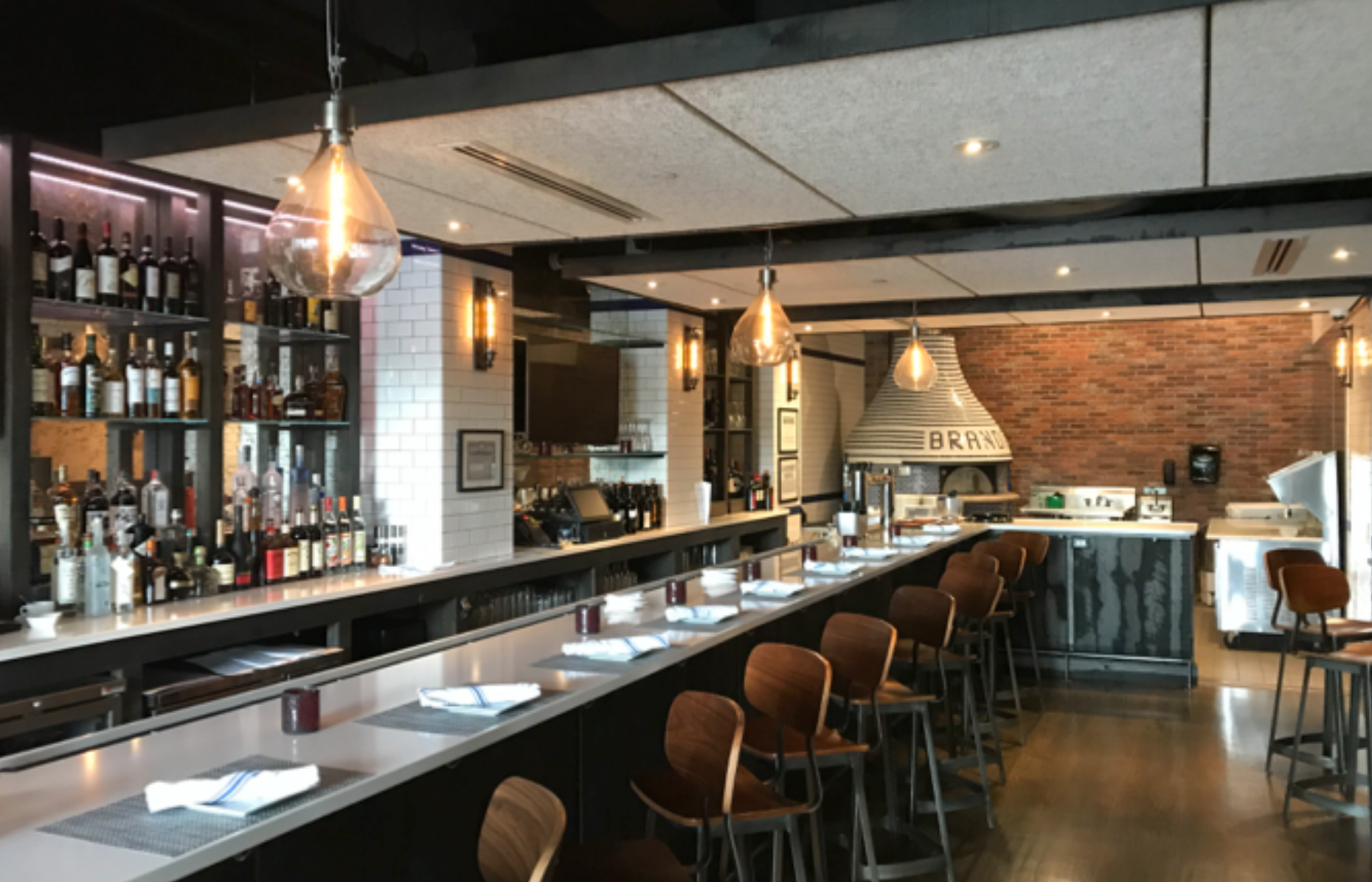 Bar interior with long counter, stools, liquor bottles, and a brick pizza oven.
