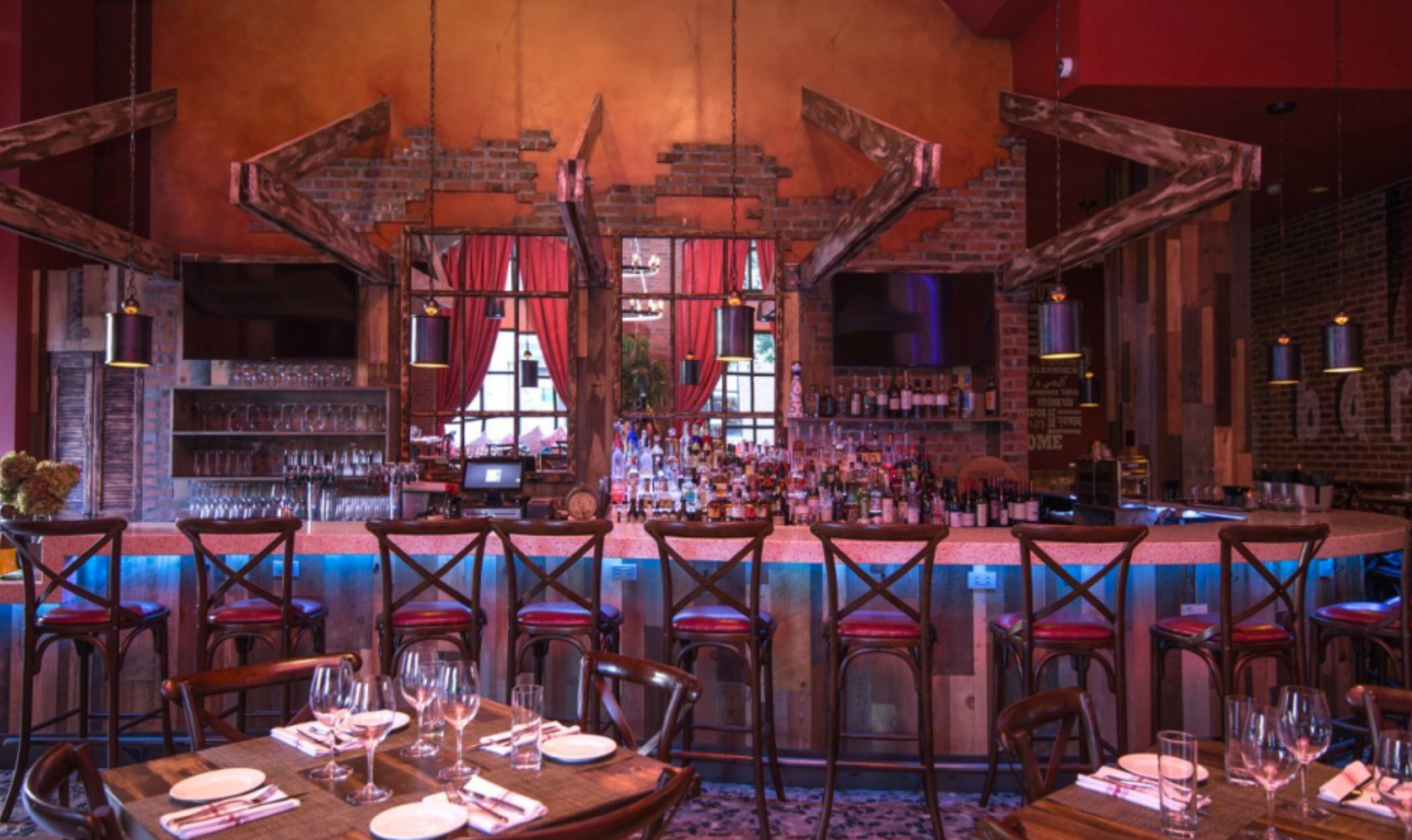 Bar with red and brown decor, lit with blue lights. Tables with place settings and chairs in foreground.