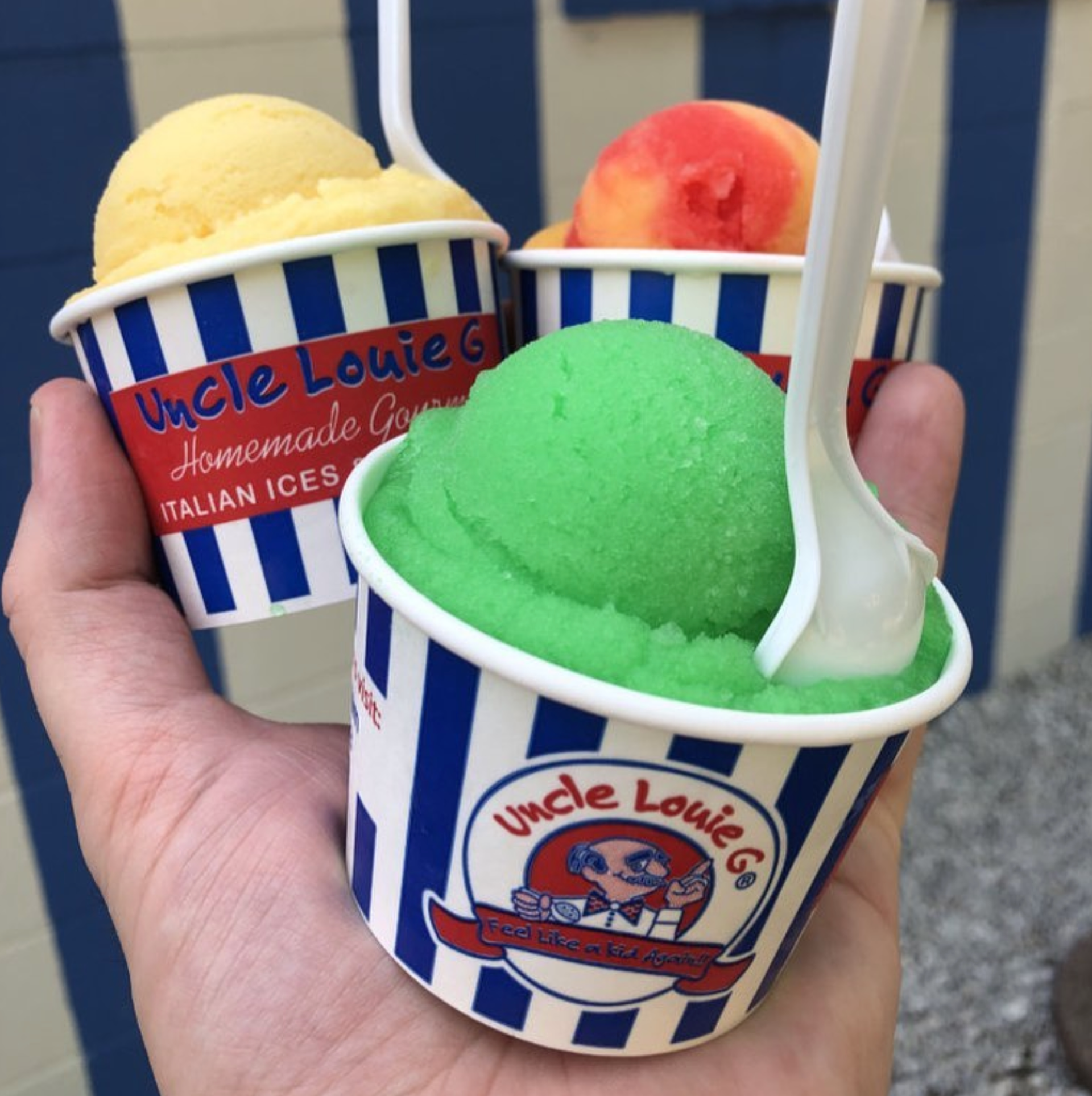 Hand holding three cups of Uncle Louie G's Italian ice: yellow, peach, and green. Cups are blue and white striped.