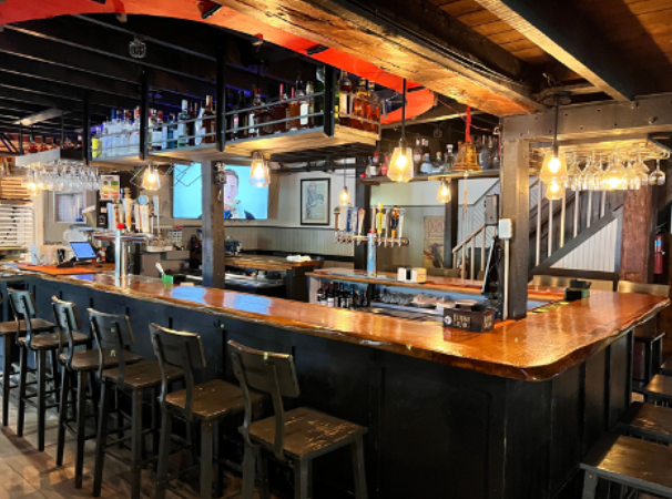 A dimly lit bar with a copper-topped counter, stools, and drinks. There are multiple TVs and exposed wooden beams.