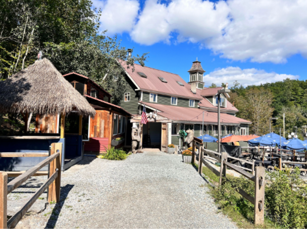 Rustic building with a thatched roof structure on the left. Stone facade and open-air seating under blue umbrellas.