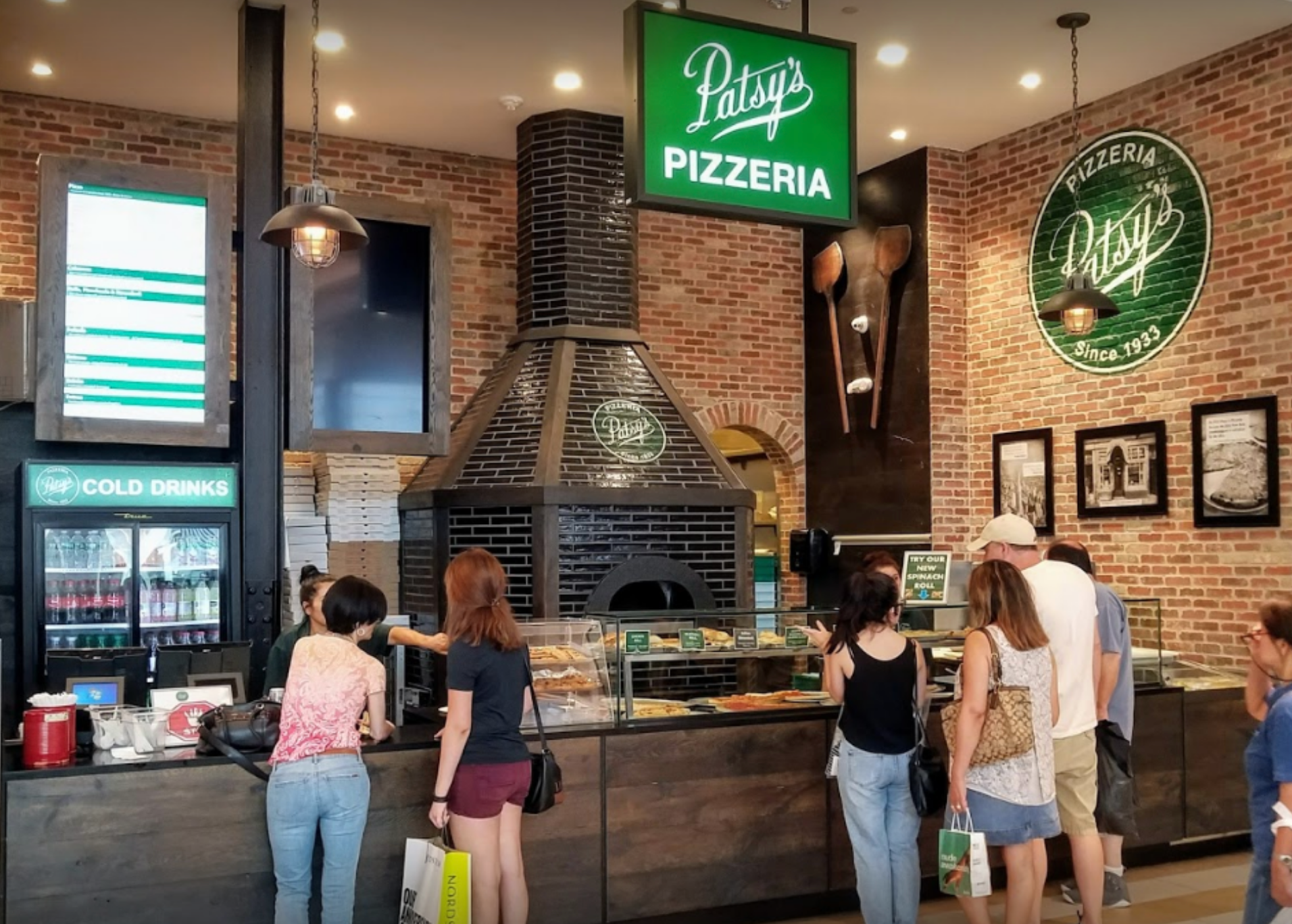 Inside Patsy's Pizzeria: Customers order at the counter, brick walls, pizza oven, green sign.