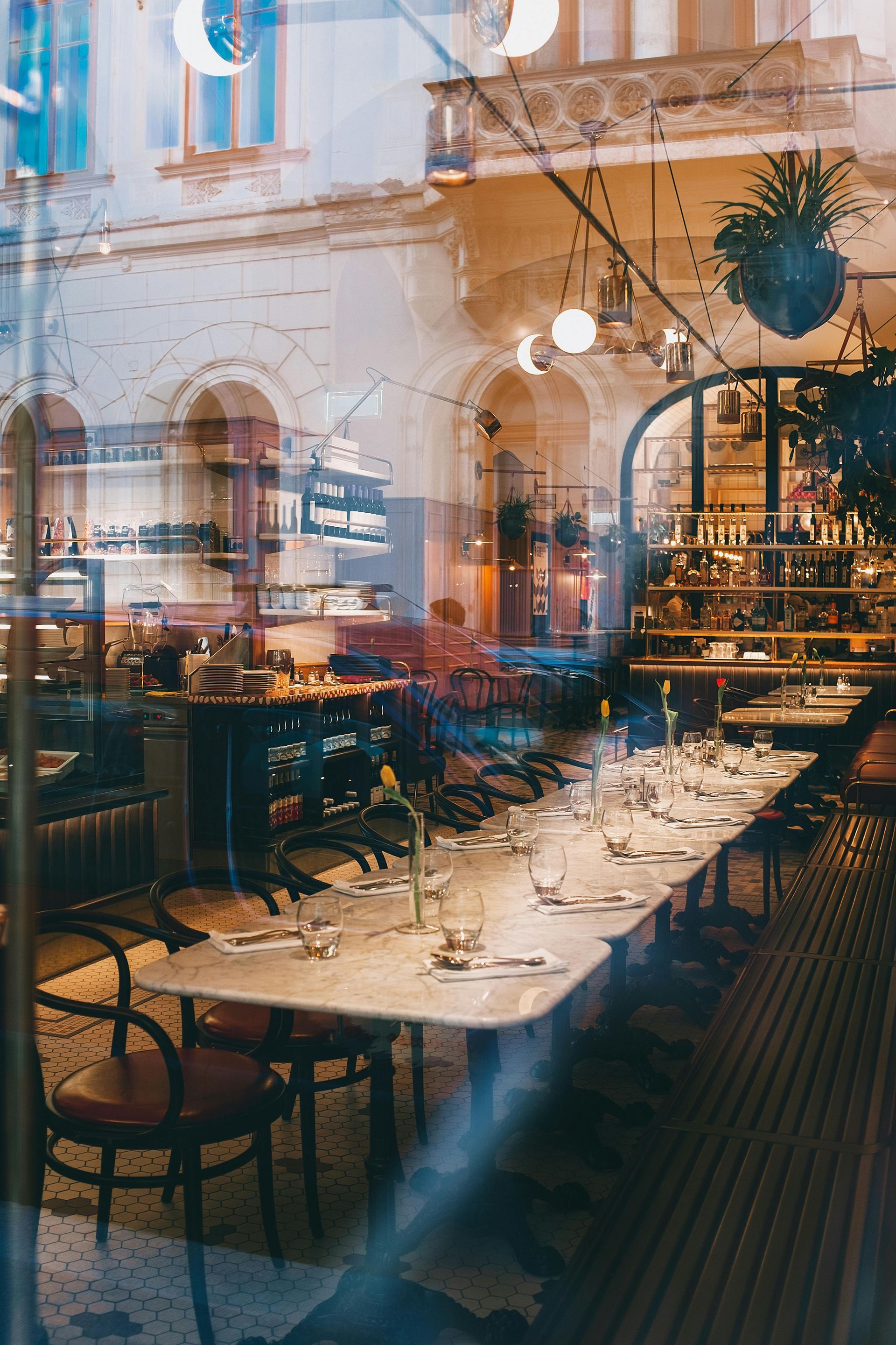 Restaurant interior, tables set for dining, seen through a reflective window. Soft lighting, hints of architecture, chairs visible.