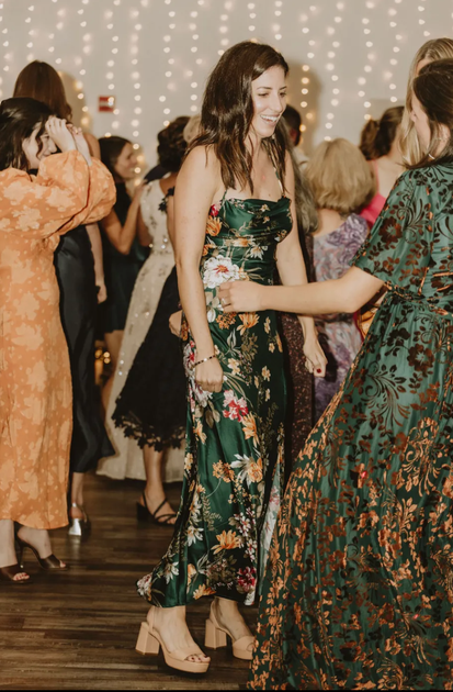 Woman in green floral dress dancing at a party. Other guests and string lights in background.