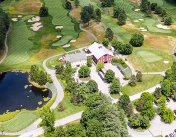 Aerial view of a golf course with a barn-like clubhouse, pond, green fairways and trees.