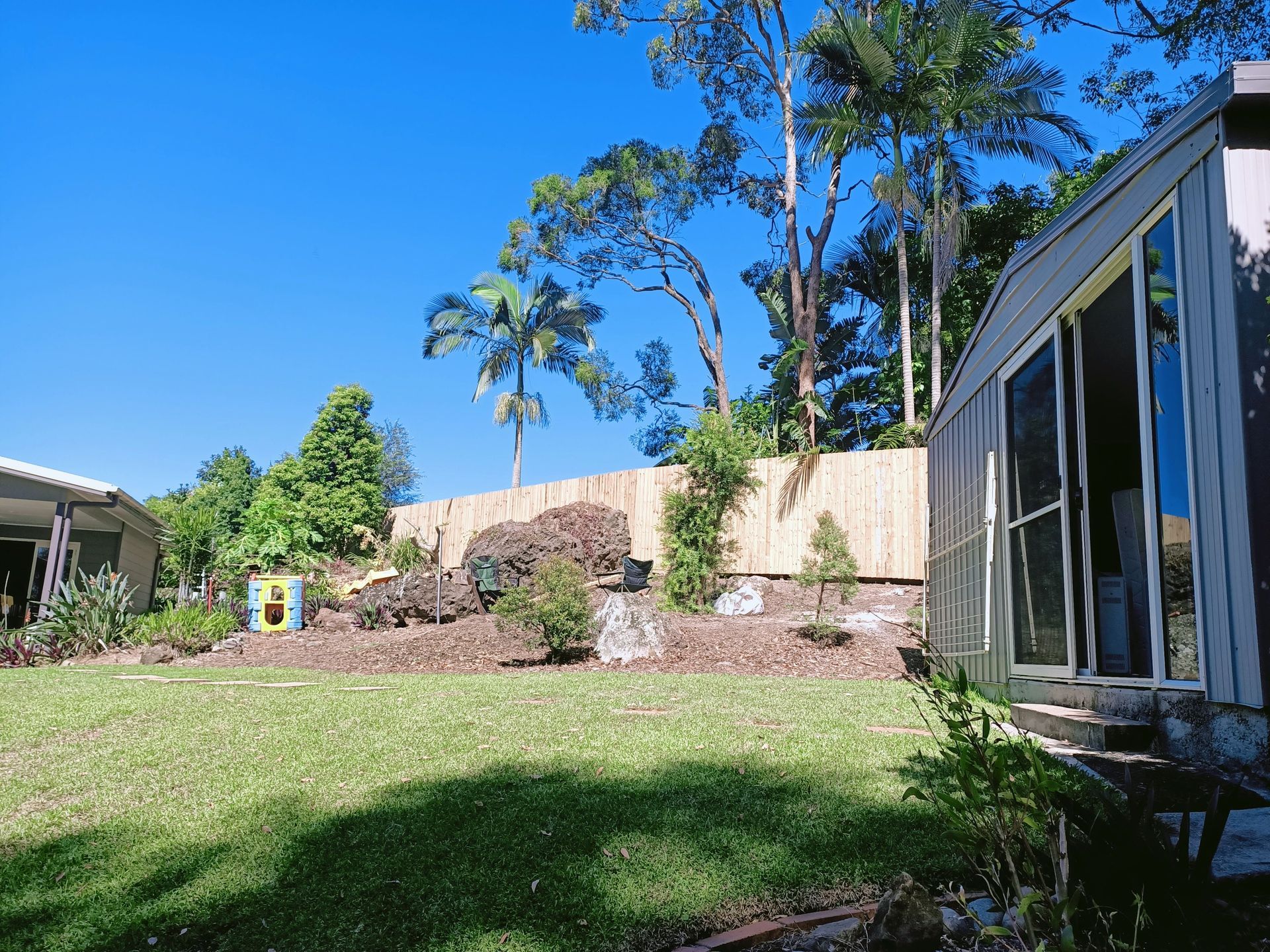 Grassy backyard with rock garden, wooden fence, trees, and house with glass sliding doors on a sunny day — Quality Fences 4 U in Gympie, QLD