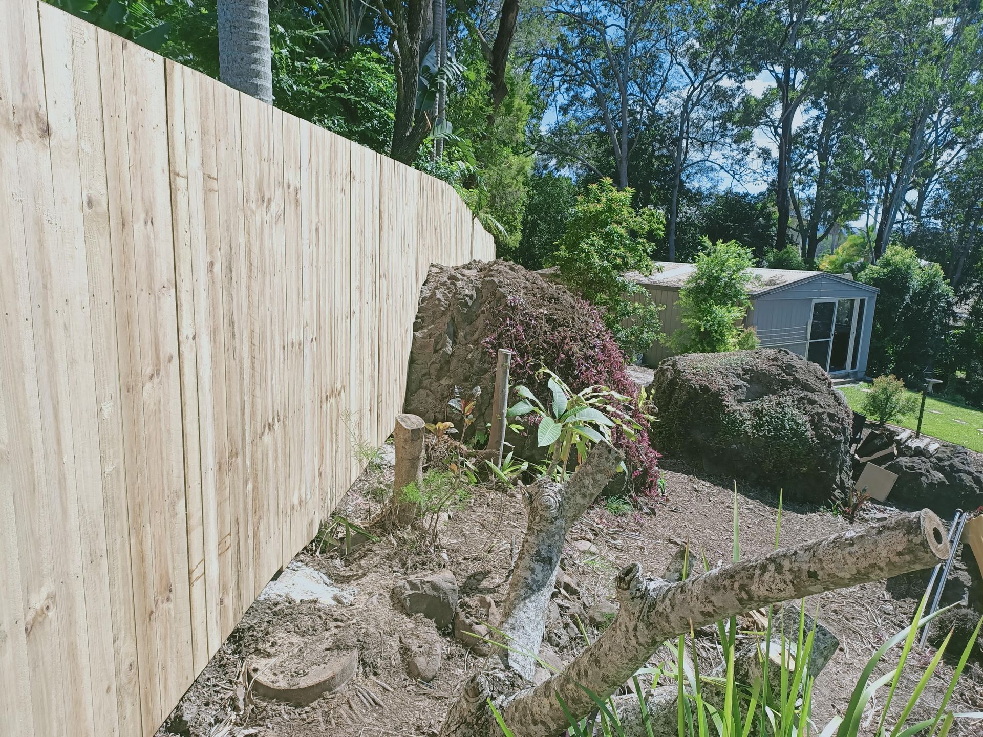 Wooden fence alongside a garden bed with pruned tree branches, rocks, and a shed in the background — Quality Fences 4 U in Gympie, QLD