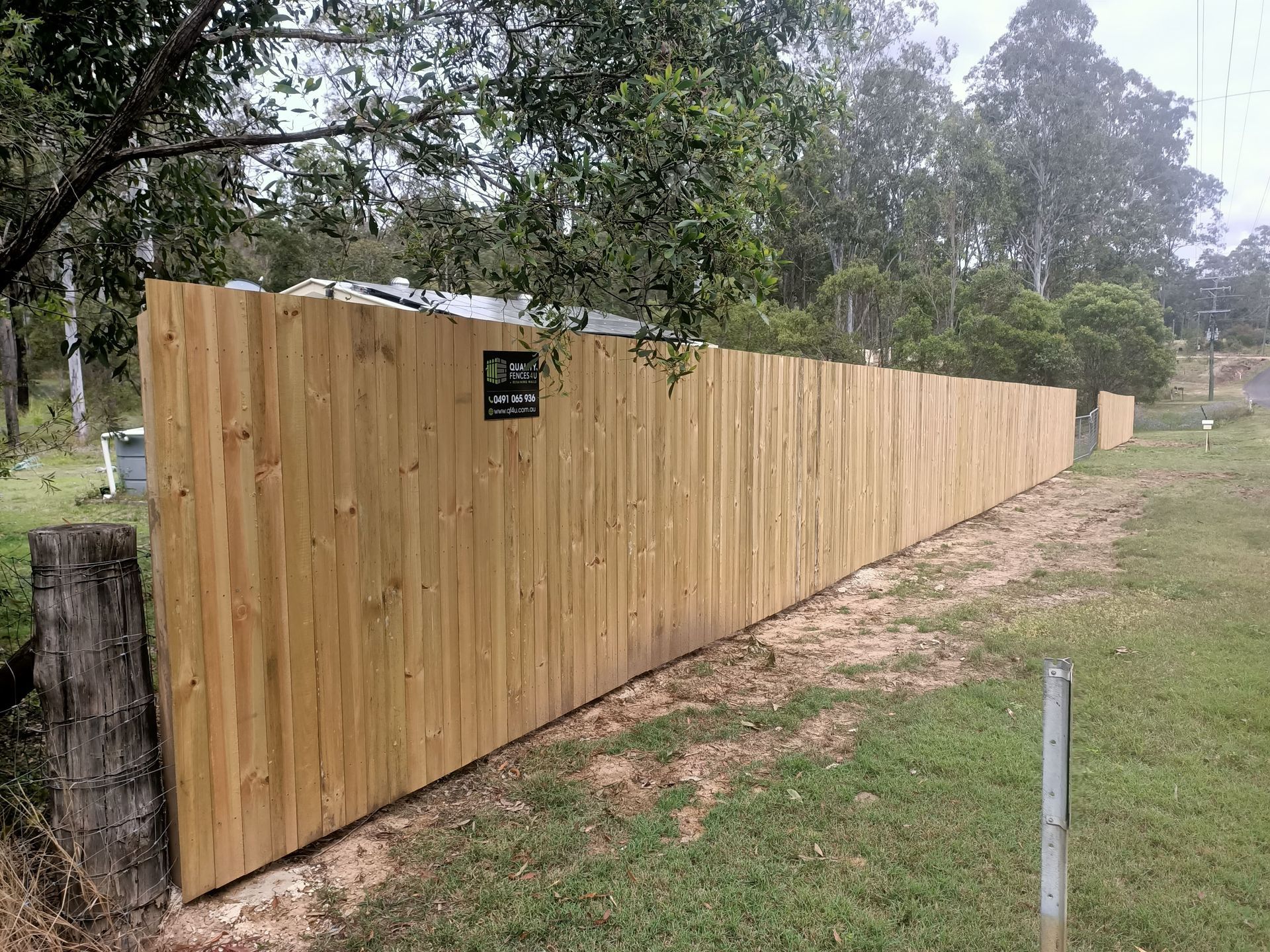 Wooden fence along a grassy area, trees in the background. A small sign is attached to the fence — Quality Fences 4 U in Gympie, QLD