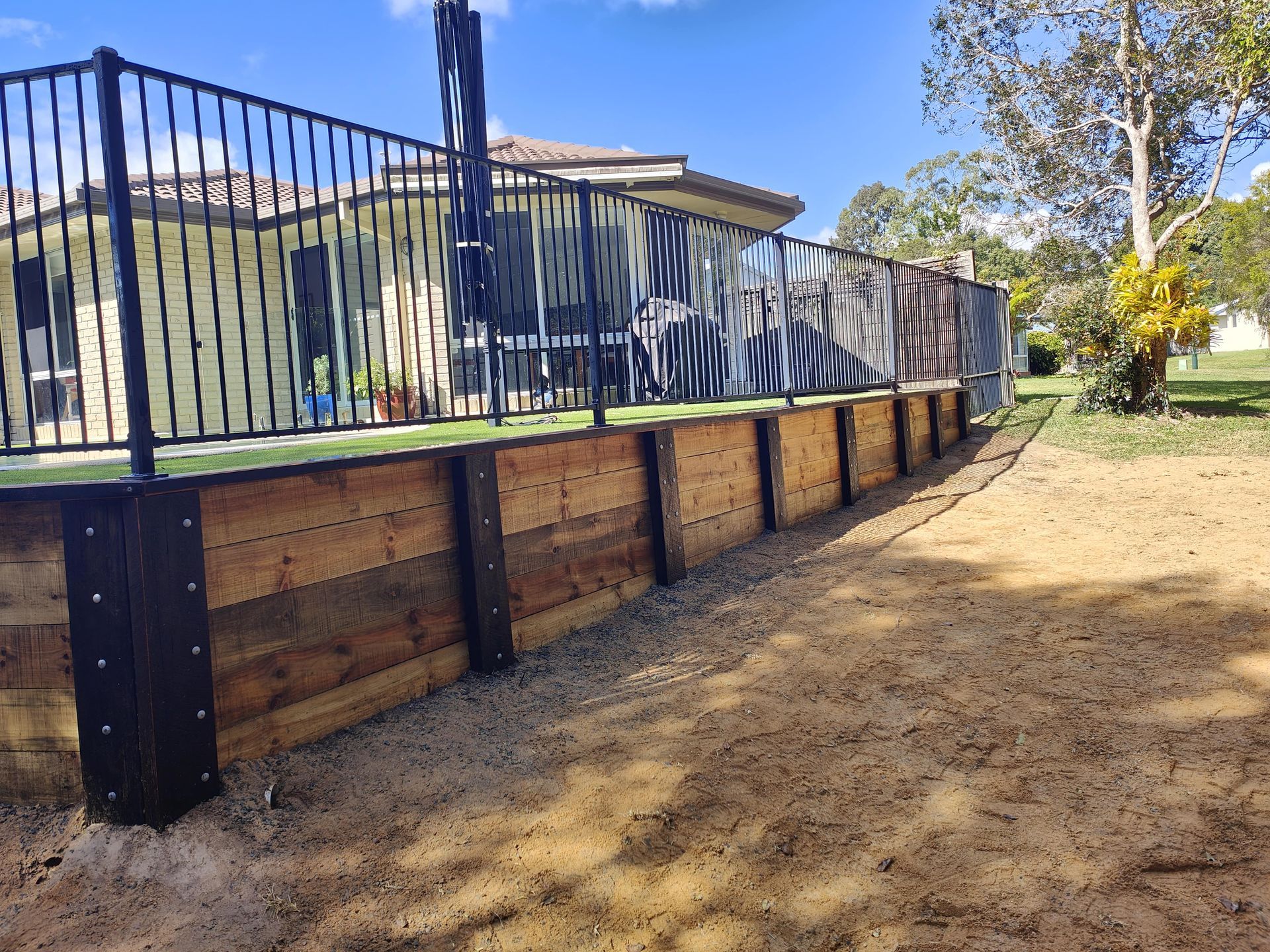 Black metal fence atop a wooden retaining wall, next to a sandy area. A house is visible in the background — Quality Fences 4 U in Gympie, QLD