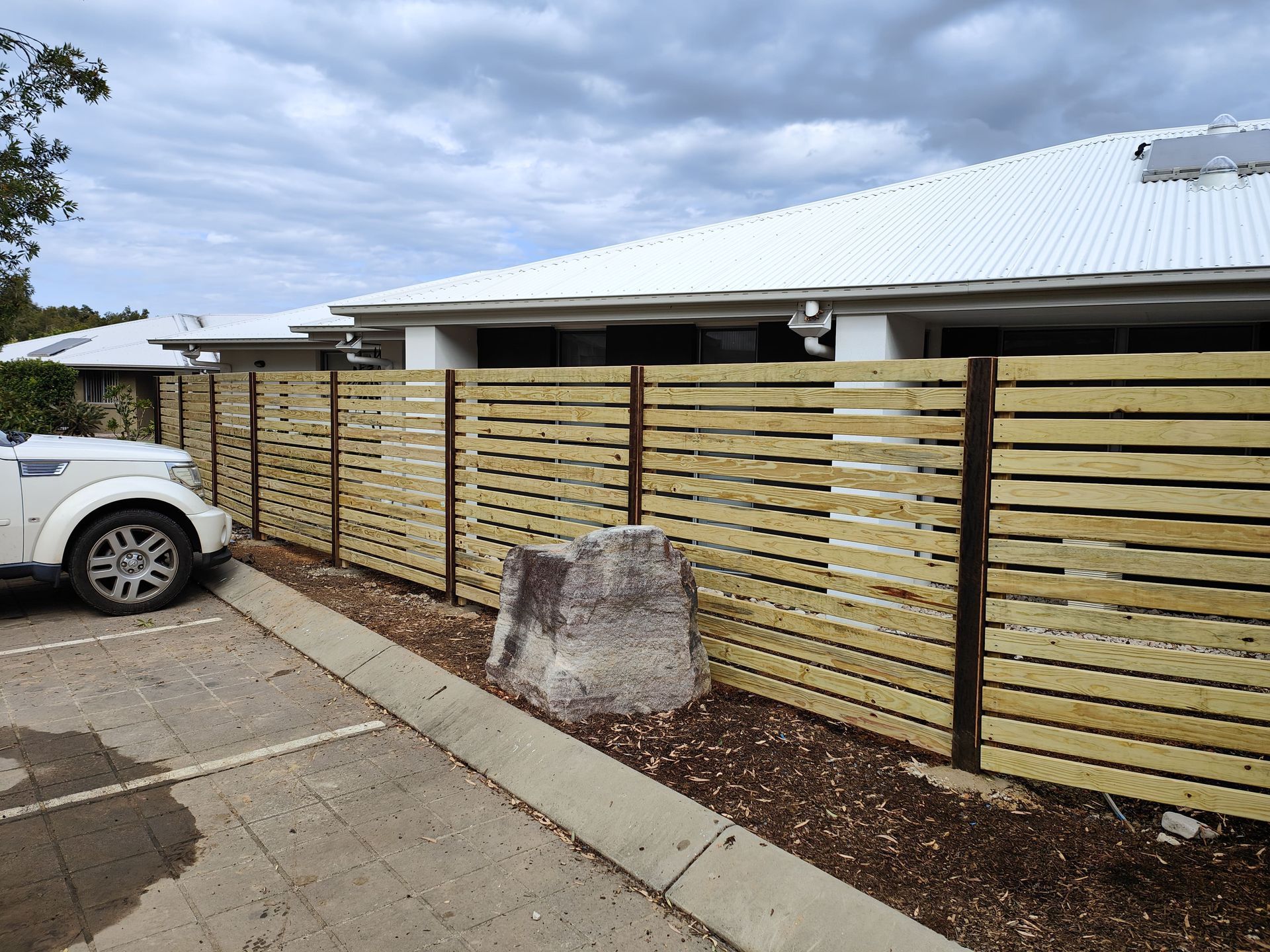 Wooden slatted fence in front of a white building with a car parked nearby on a paved area, cloudy sky — Quality Fences 4 U in Gympie, QLD