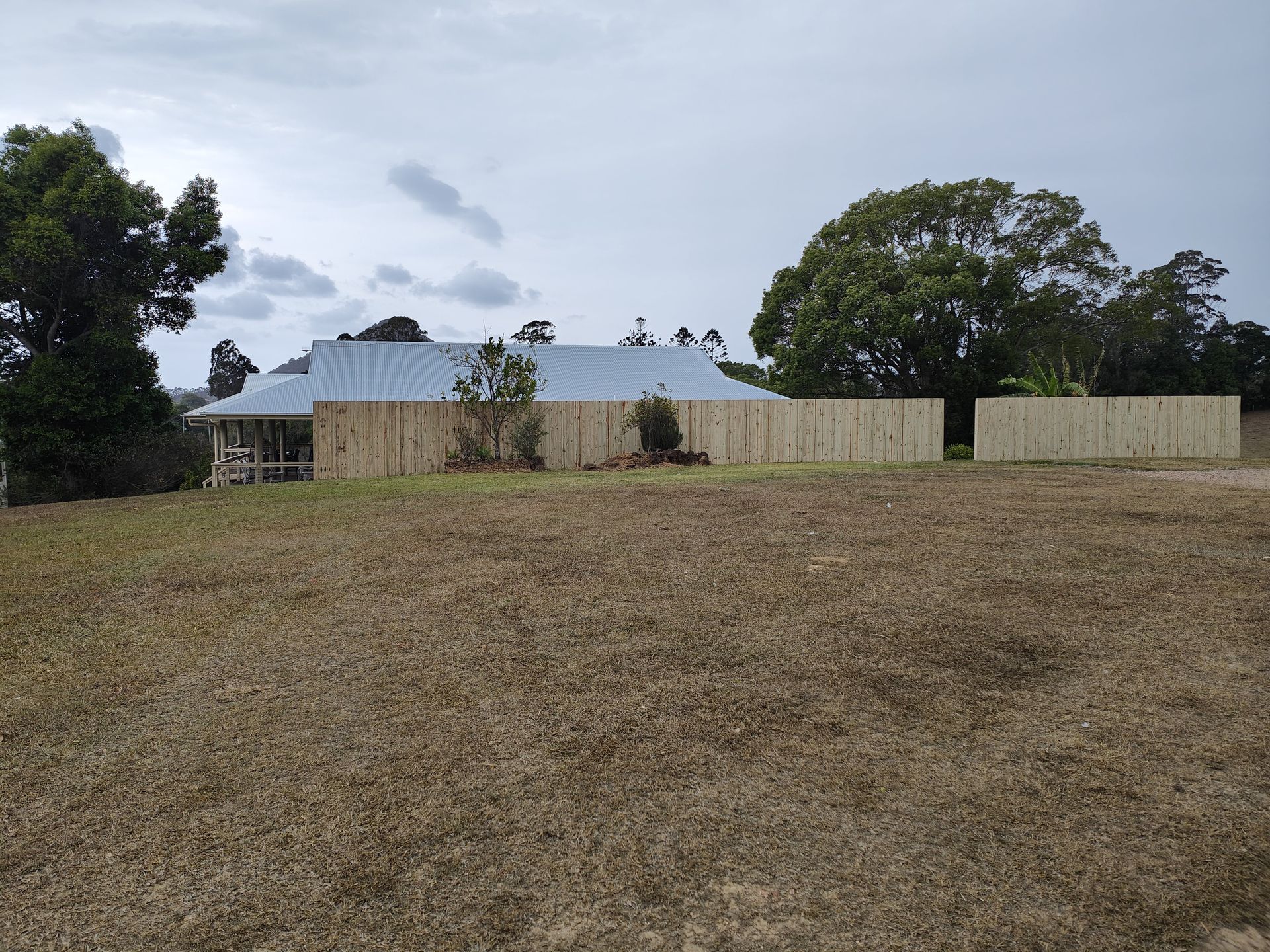 A house with a silver roof behind a light wood fence and dry grass under a cloudy sky — Quality Fences 4 U in Gympie, QLD