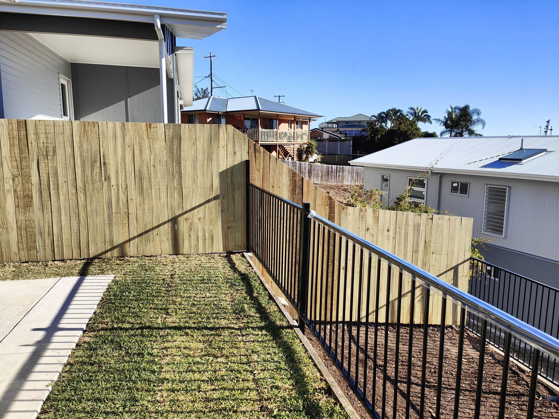 Wooden fence, black railing, and grass yard with houses in the background under a blue sky — Quality Fences 4 U in Gympie, QLD