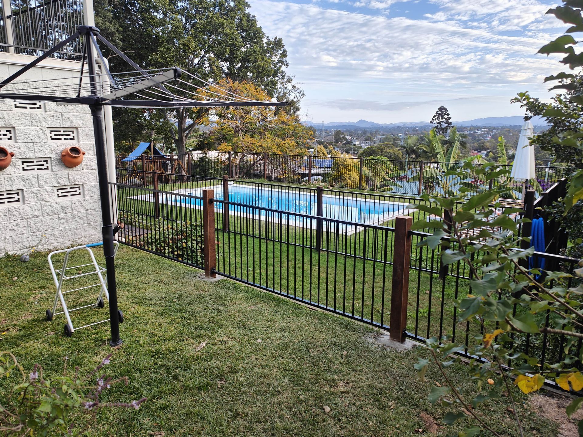Lush green yard with a black fence surrounds a blue swimming pool, overlooking a town under a cloudy sky — Quality Fences 4 U in Gympie, QLD