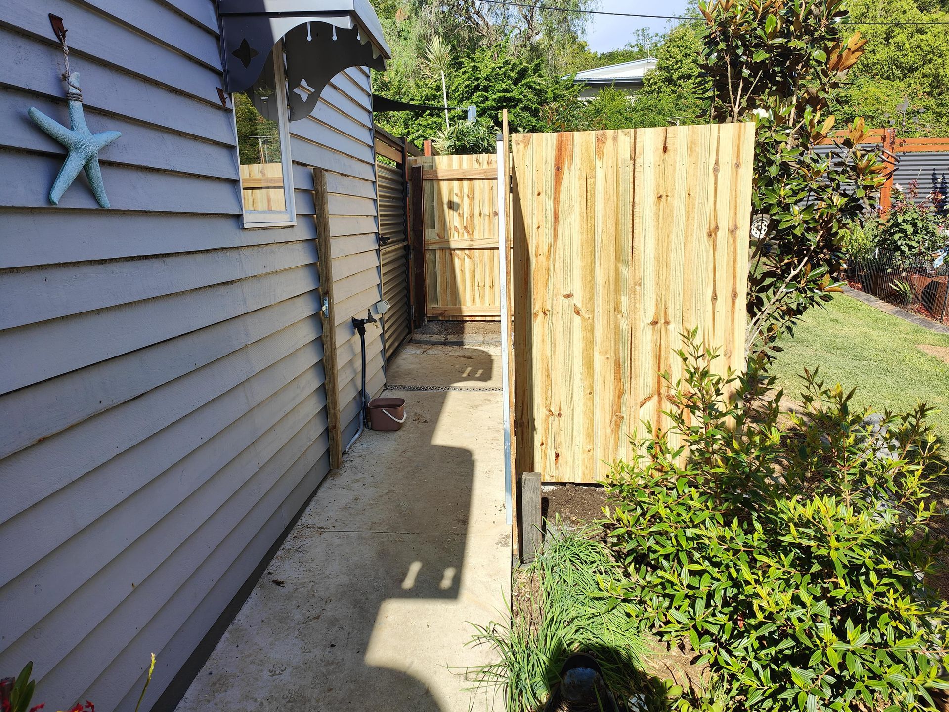 A narrow concrete walkway between a light blue building and wooden fence, with green foliage on the right — Quality Fences 4 U in Gympie, QLD