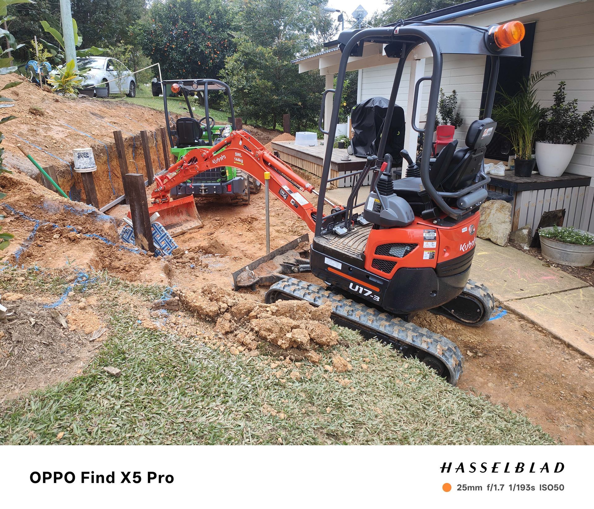 An orange mini-excavator digging a trench near a house; another excavator is in the background — Quality Fences 4 U in Gympie, QLD