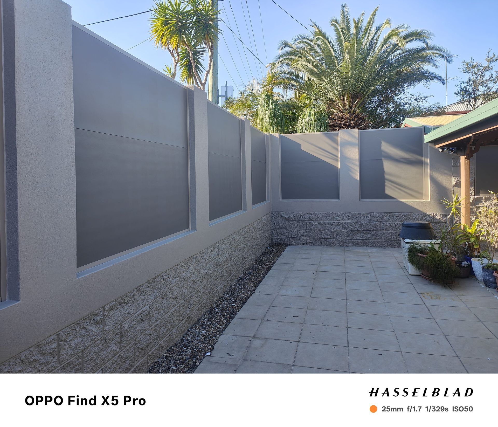 Gray concrete wall with dark gray panels, over a gravel base, next to a paved patio, with a few plants and trees in the background — Quality Fences 4 U in Gympie, QLD