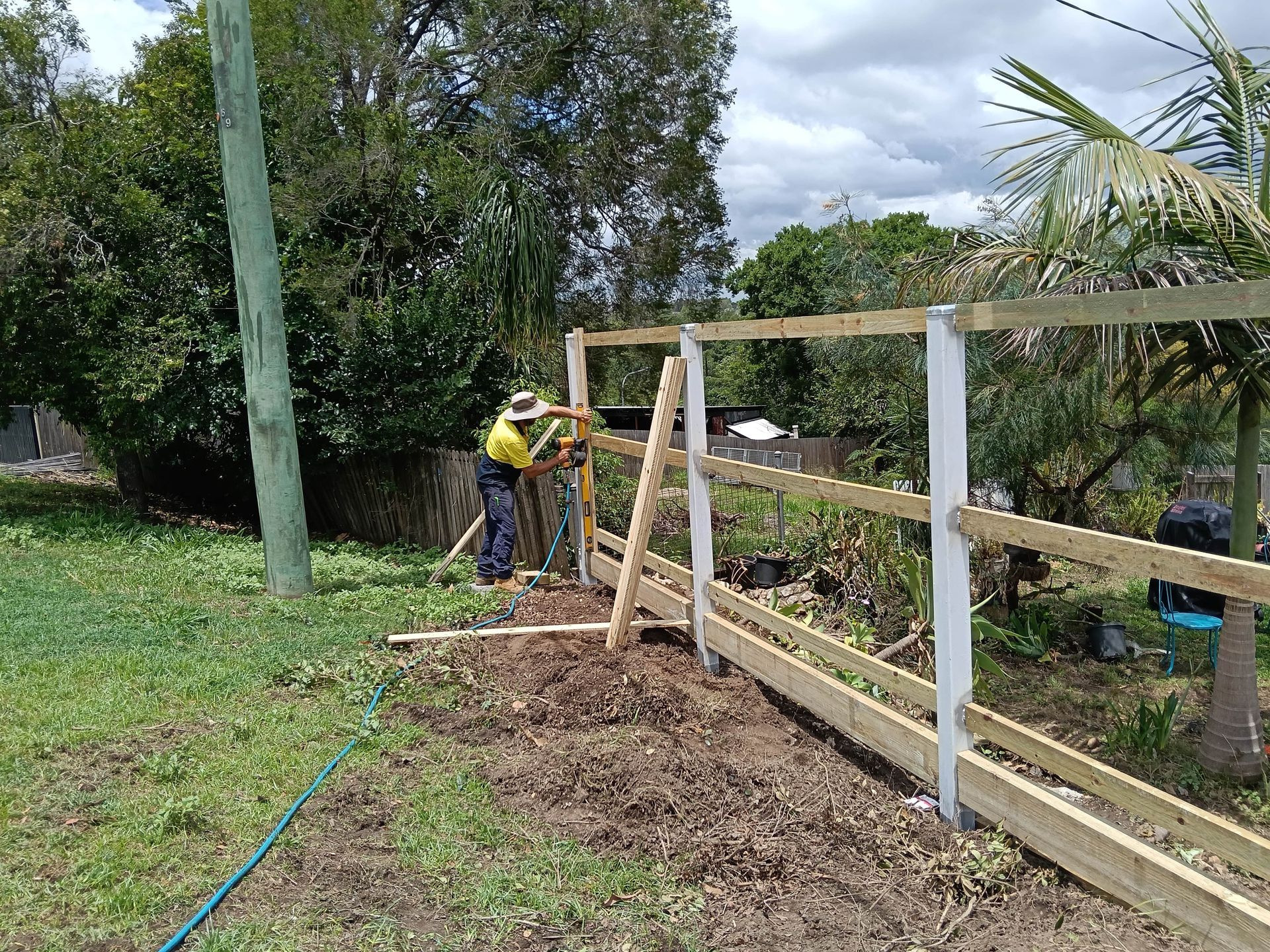 Person in a yellow shirt constructing a wooden fence in a yard with green grass and trees — Quality Fences 4 U in Gympie, QLD