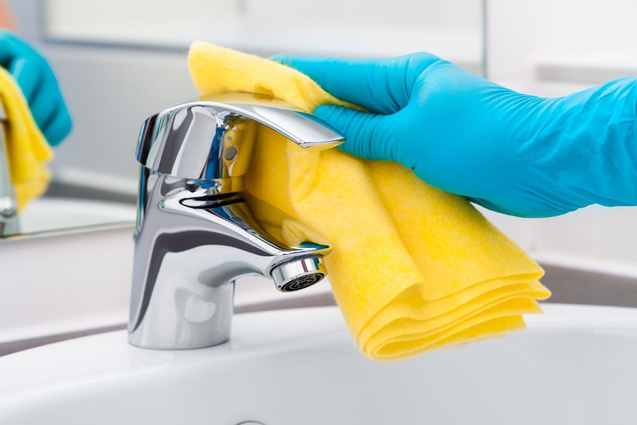 A person wearing blue gloves is cleaning a sink faucet with a yellow cloth.