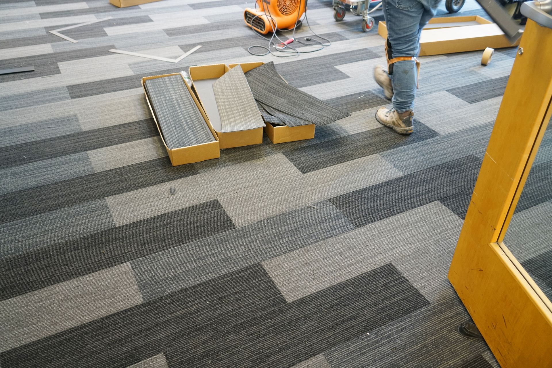 Person standing on newly installed carpet; samples on cardboard, floor fan in background.