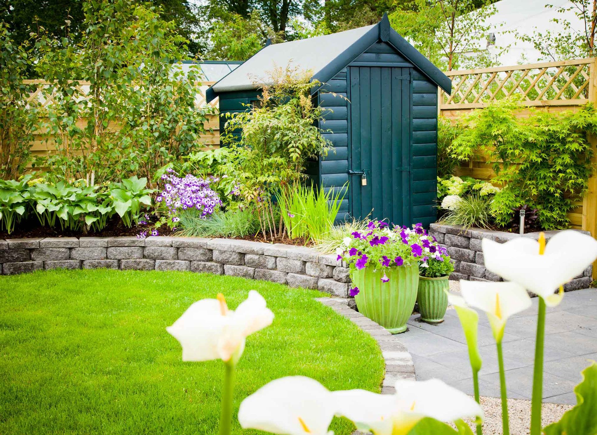 Green shed in a garden with lawn, flowers, stone wall, and potted plants.