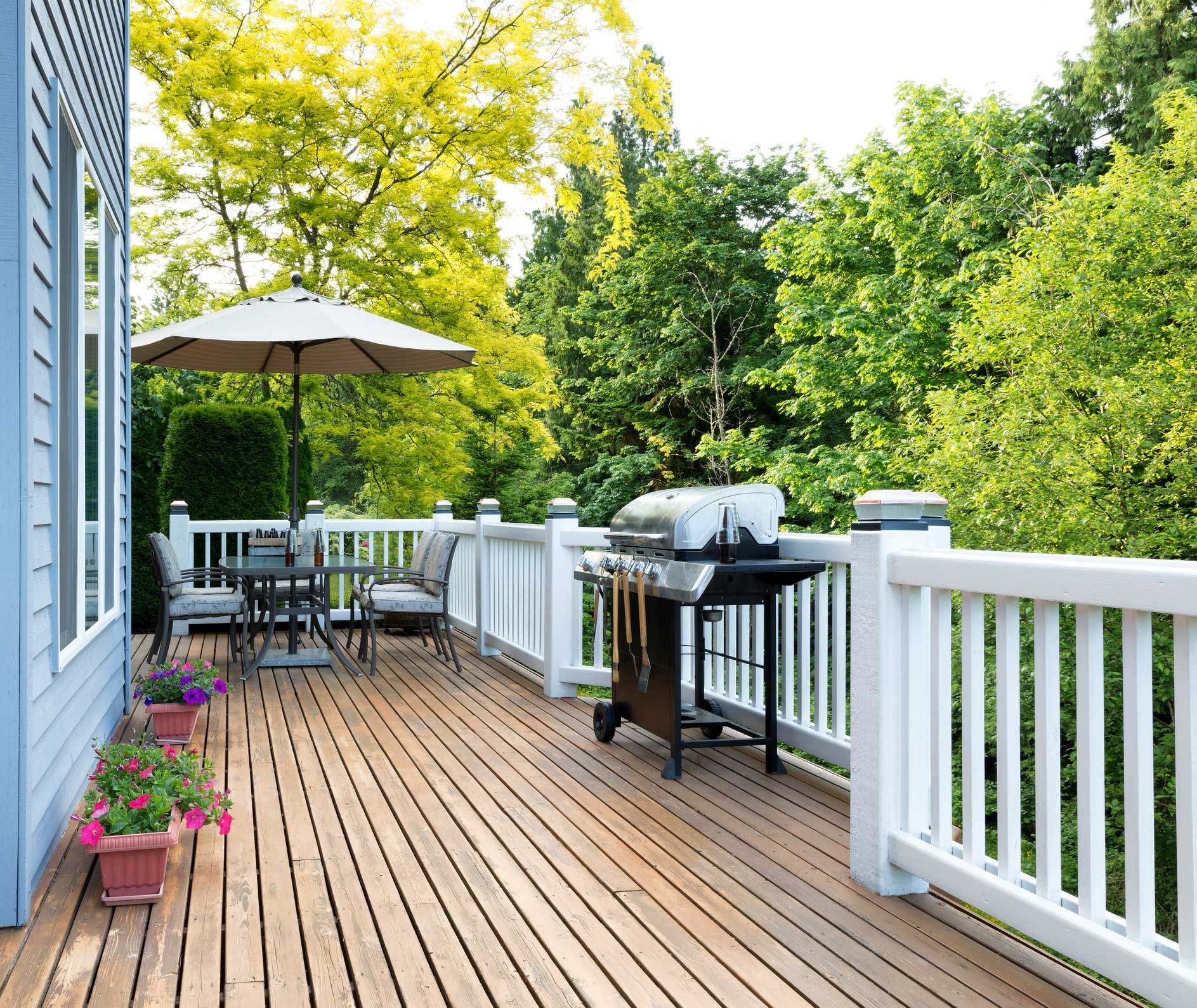 Wooden deck with a grill and patio furniture, surrounded by lush green trees.