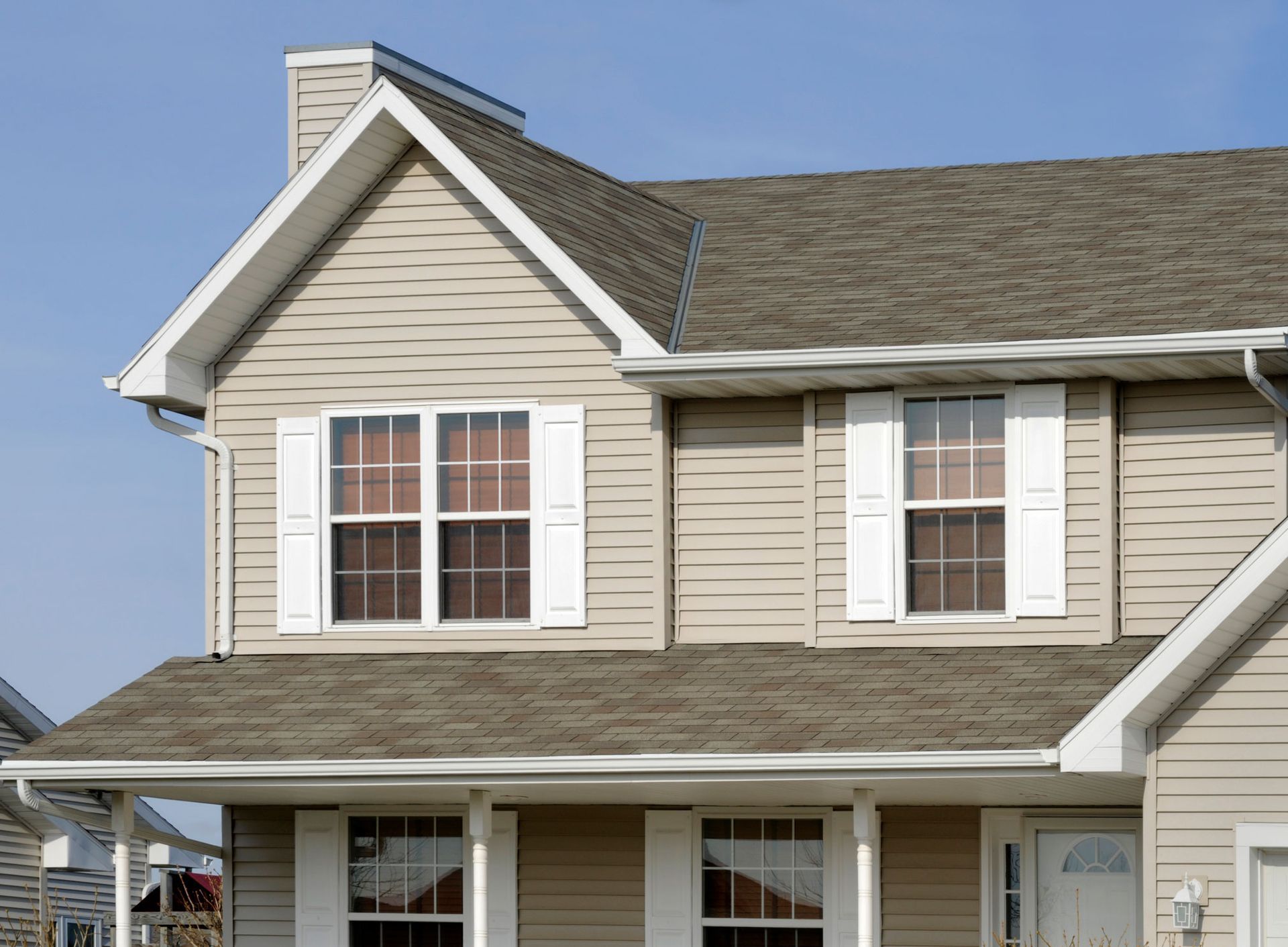 Two-story beige house with white trim, shutters, and brown roof against a blue sky.