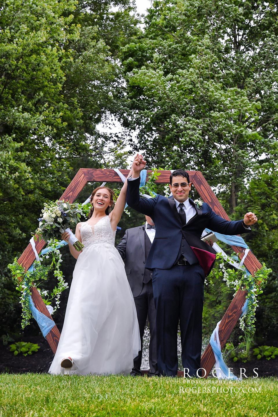 A bride and groom are holding their hands in the air in front of a wooden arc at Woodwinds  wedding photographed by Rogers Photography in Connecticut.