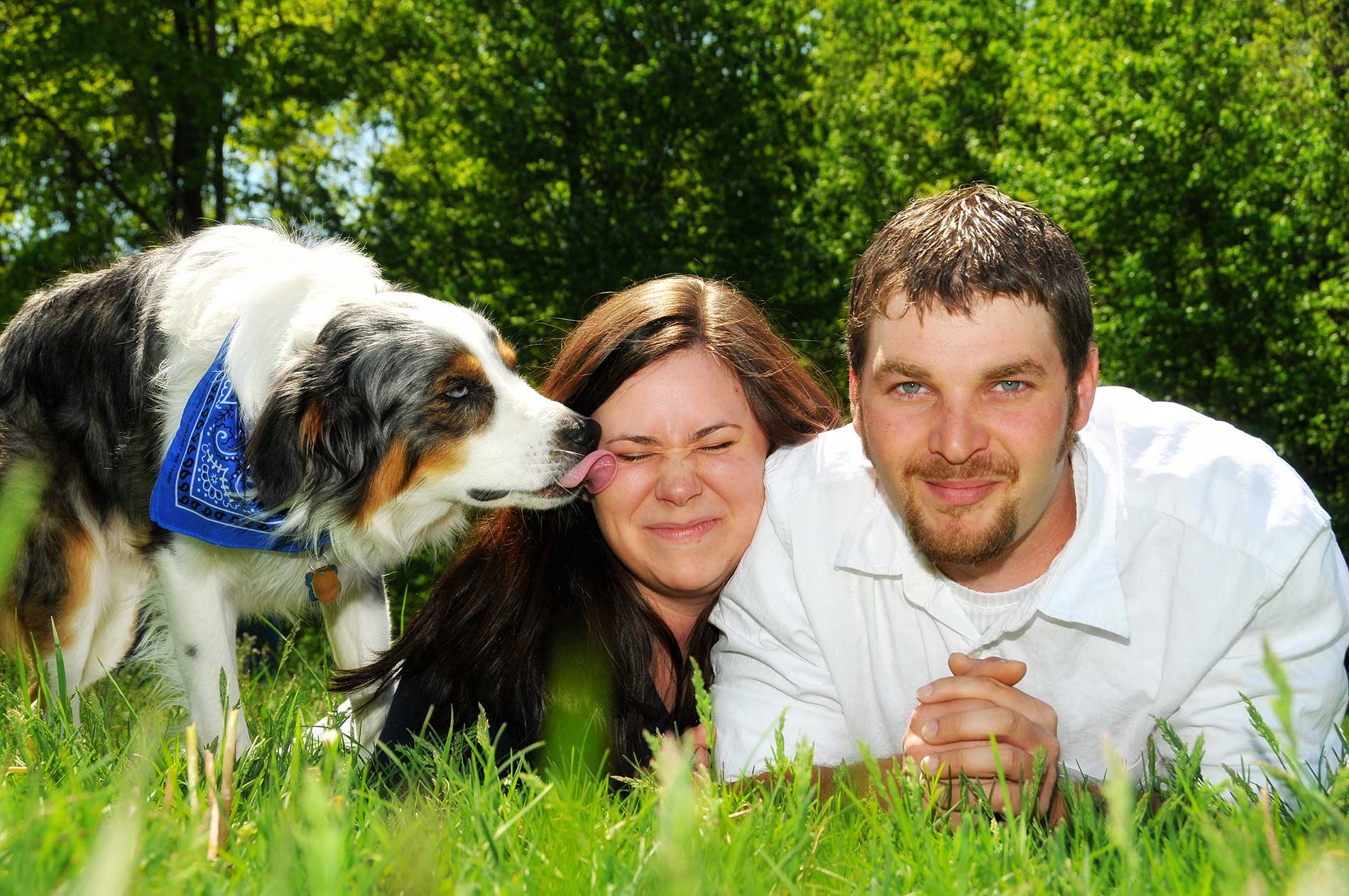 An engagement photo of man and woman laying in the grass with a dog kissing the woman by Rogers Photography