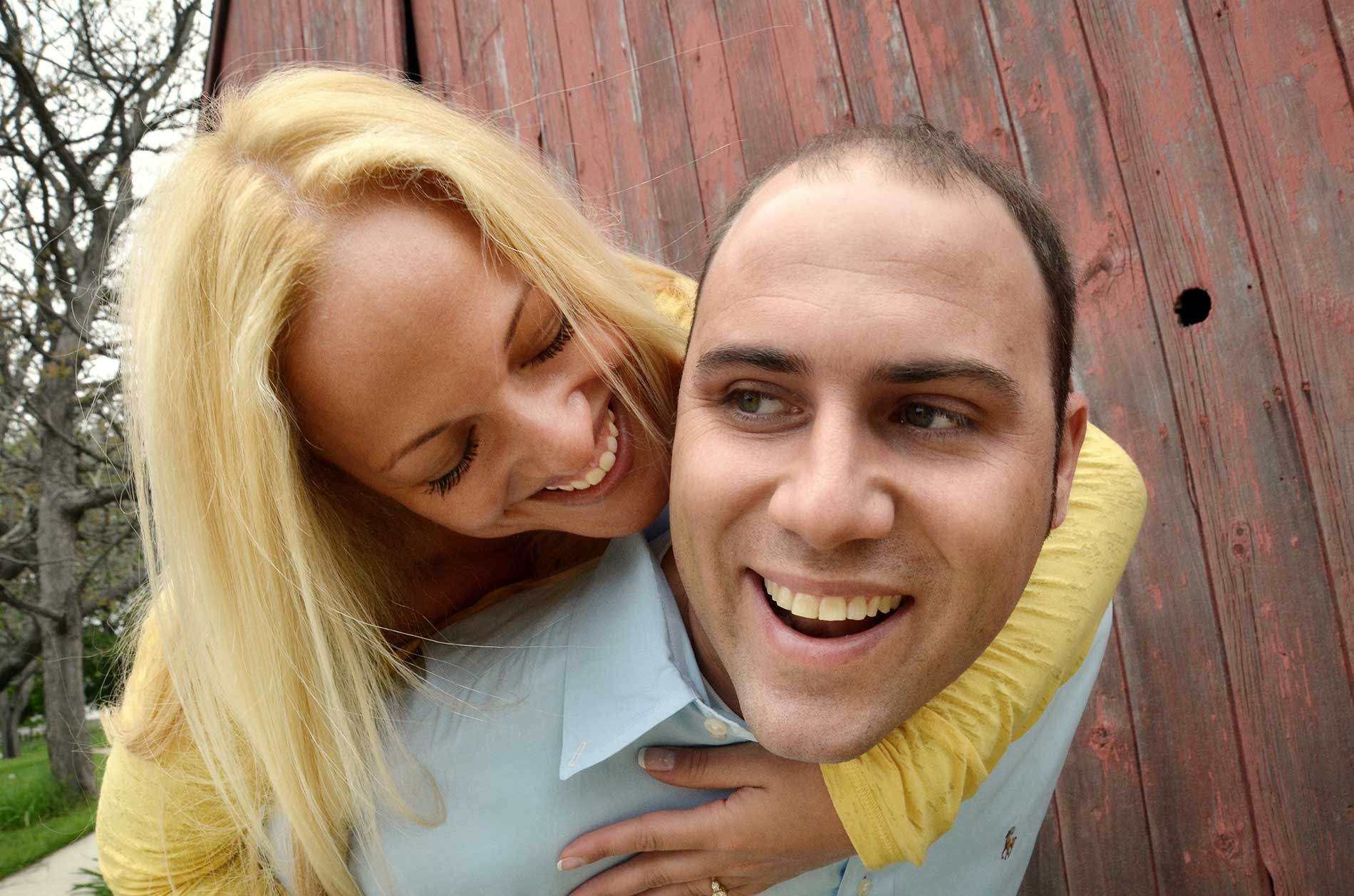 A woman is giving a man a piggyback ride and by a barn in Guilford, CT for an engagement photo by Rogers Photography