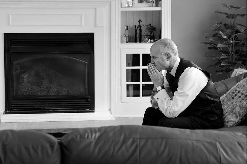 father of the bride sits on a couch with his hands folded in contemplation in front of a vase of flowers after major photo retouching