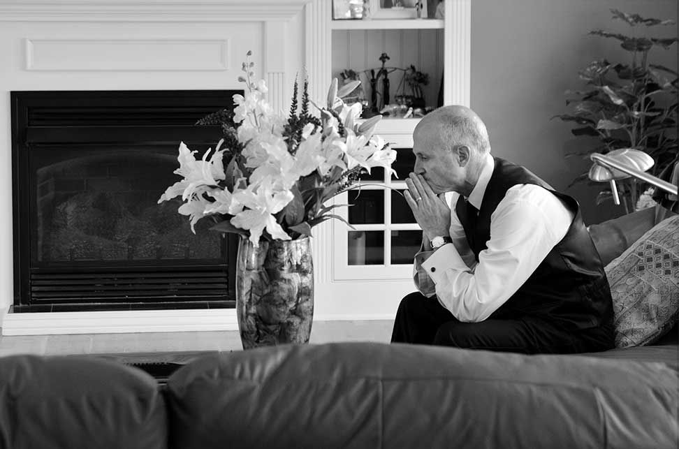 father of the bride sits on a couch with his hands folded in contemplation in front of a vase of flowers before major photo retouching
