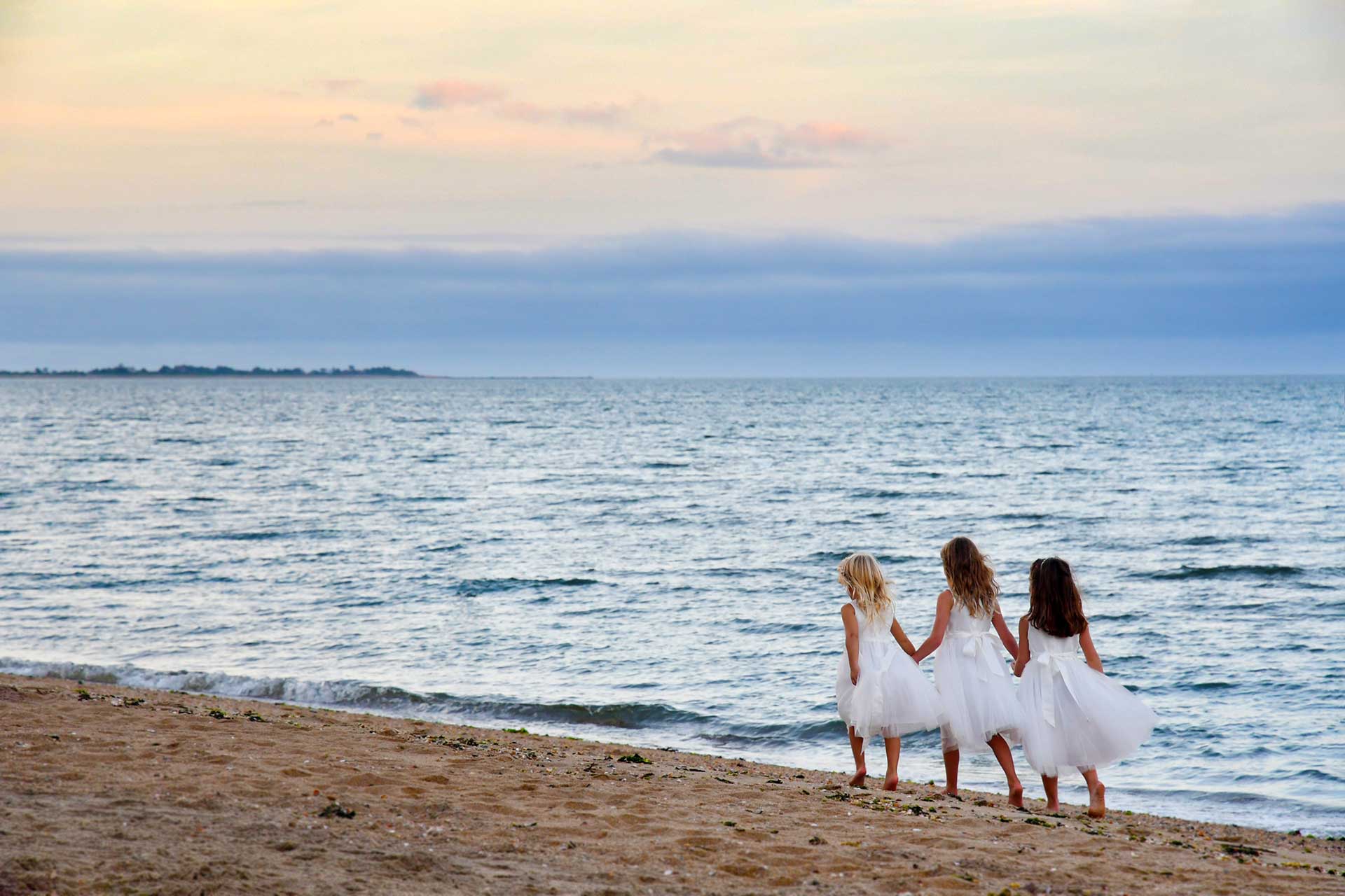 Three flower girls in white dresses are standing on a beach looking at the ocean wedding photographed by Rogers Photography in Connecticut.