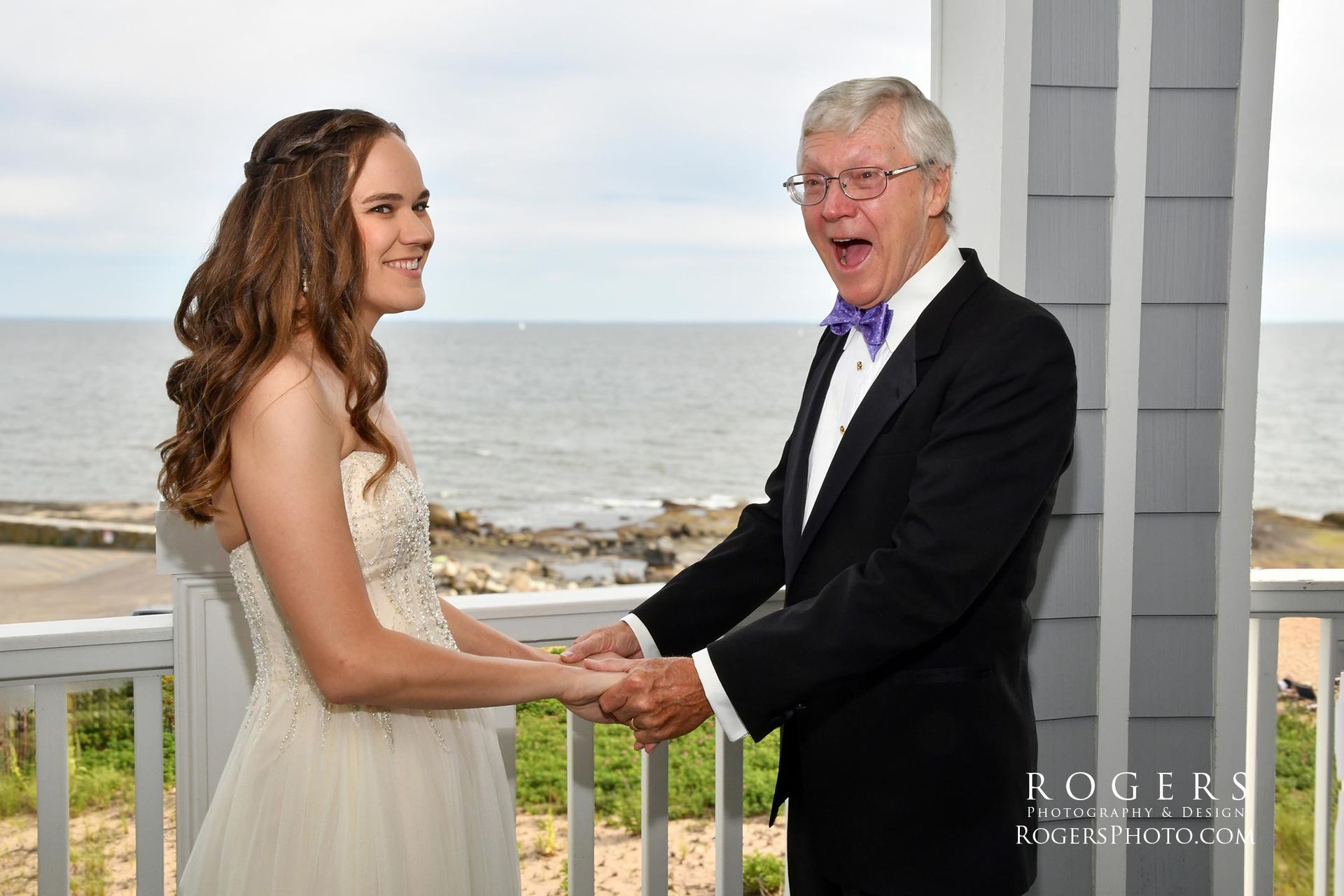 A father in a tuxedo and a bride in a wedding dress are holding hands on a balcony overlooking the ocean at Madison Beach Hotel wedding photographed by Rogers Photography in Connecticut.