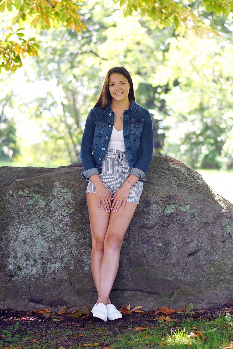 A senior portrait young girl is leaning against a large rock in a park by Rogers Photography.