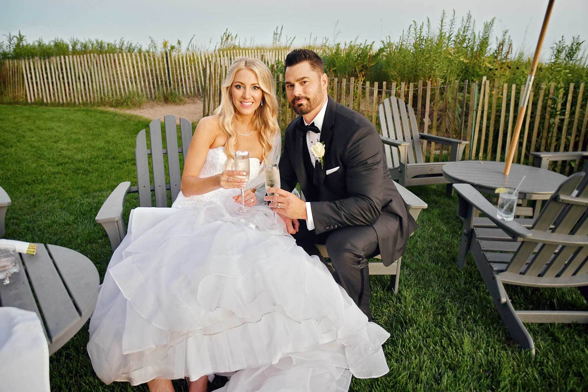 A bride and groom are sitting in lawn chairs holding champagne glasses by the beach at Madison Beach Hotel wedding photographed by Rogers Photography in Connecticut.