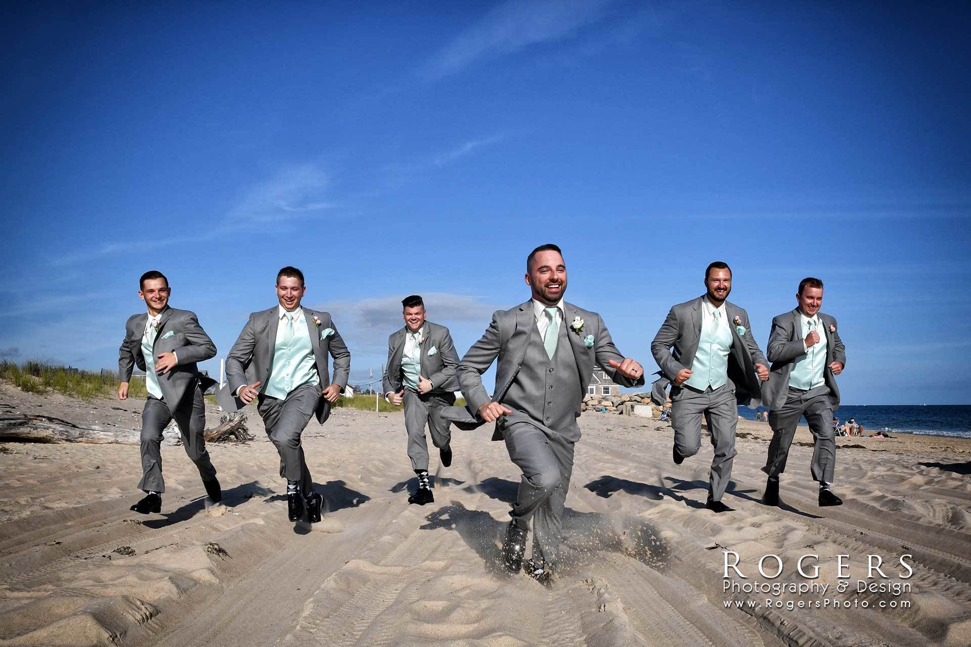 A groom and groosmen in suits and ties are running on a sandy beach wedding photographed by Rogers Photography in Connecticut.