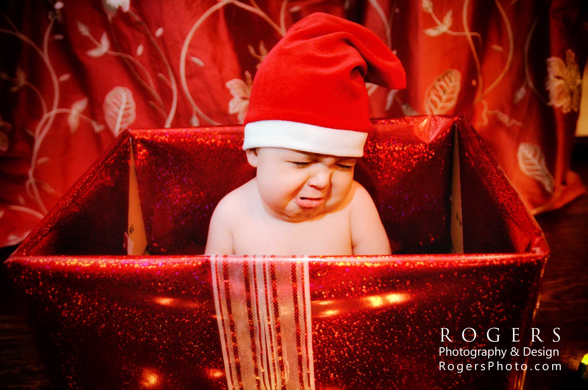 An unhappy baby wearing a santa hat is in a red box for a Christmas Card by Rogers Photography.