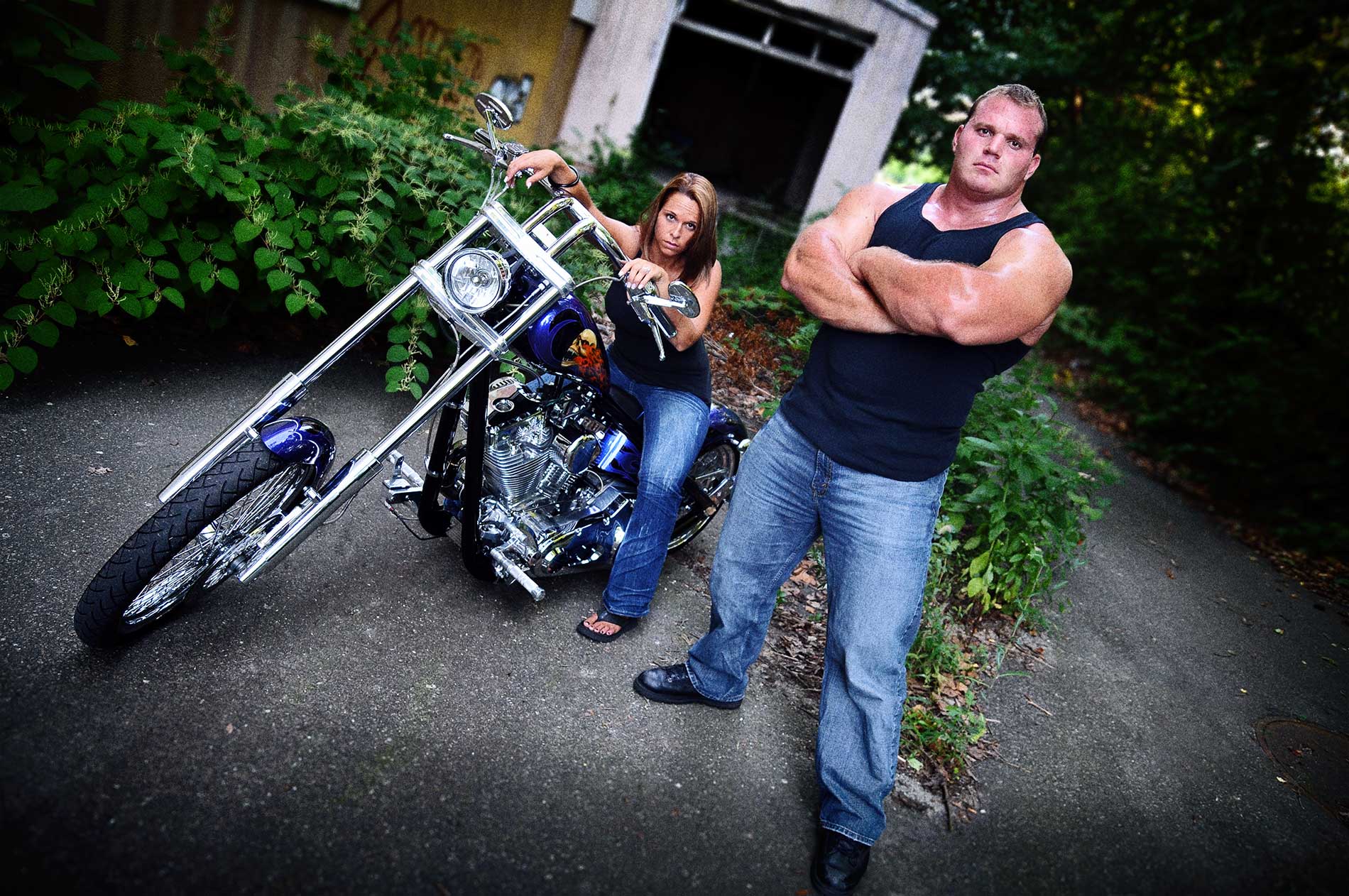 A muscle man and a woman standing next to a motorcycle for an engagement sitting by Rogers Photography.