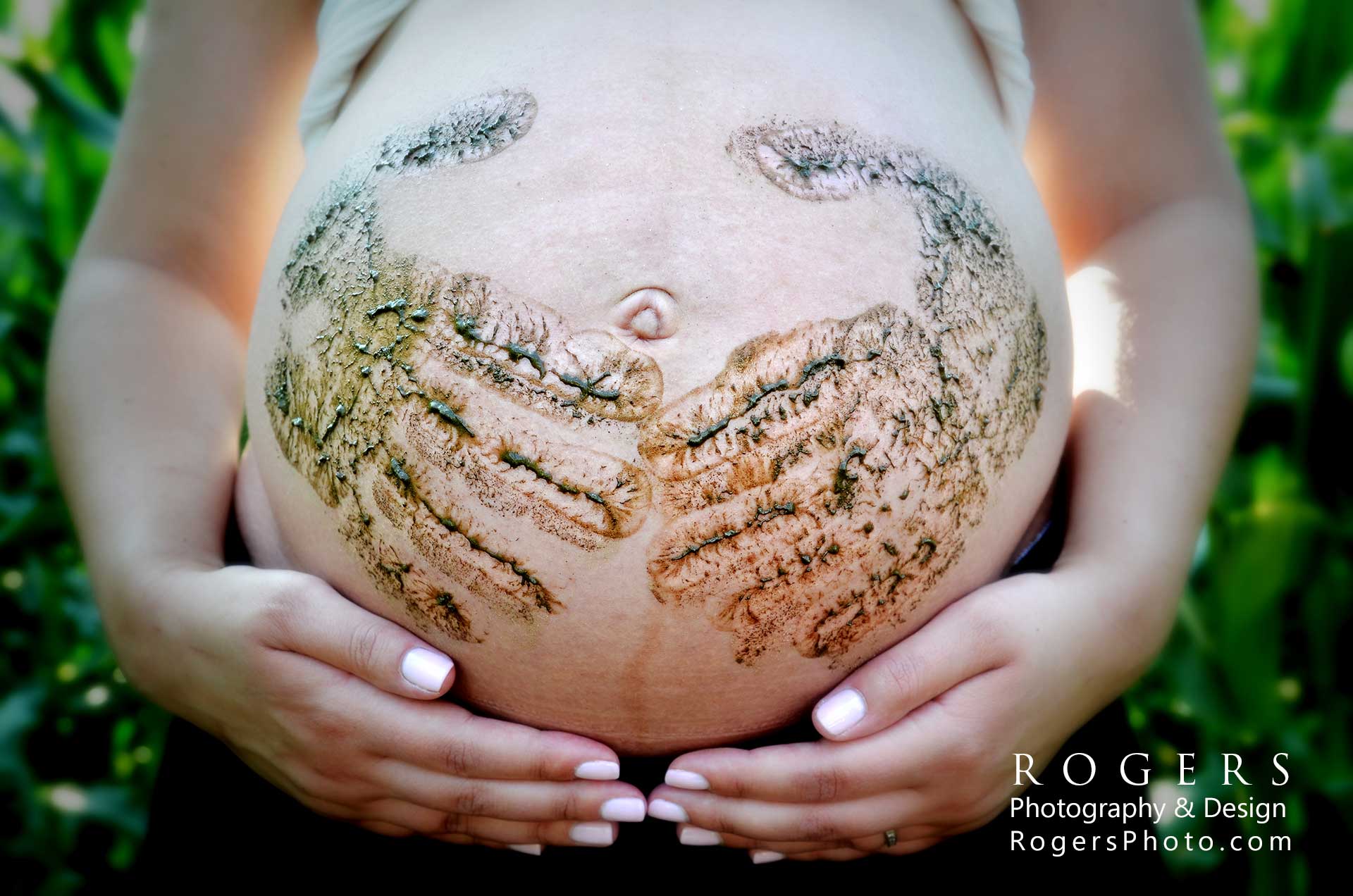 A pregnant woman is holding her belly with muddy hand prints on it by Rogers Photography.
