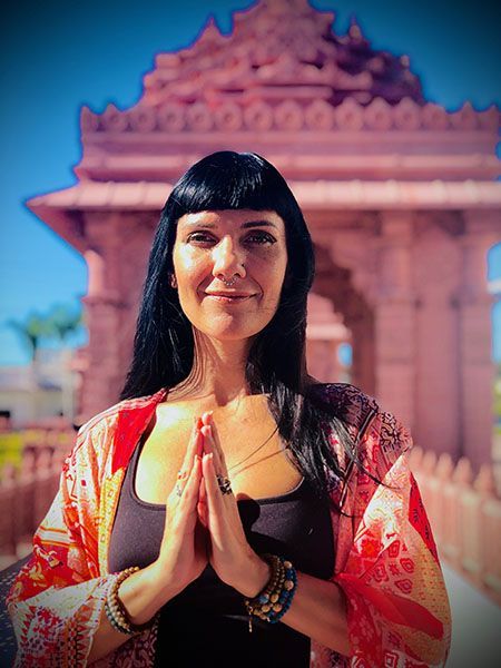 Woman with long black hair stands in prayer pose before a temple, wearing a red patterned shawl.