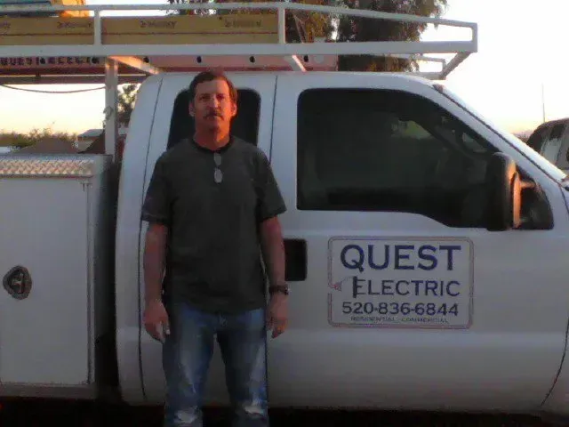 Man standing beside a white QUEST Electric service truck with ladder racks at sunset