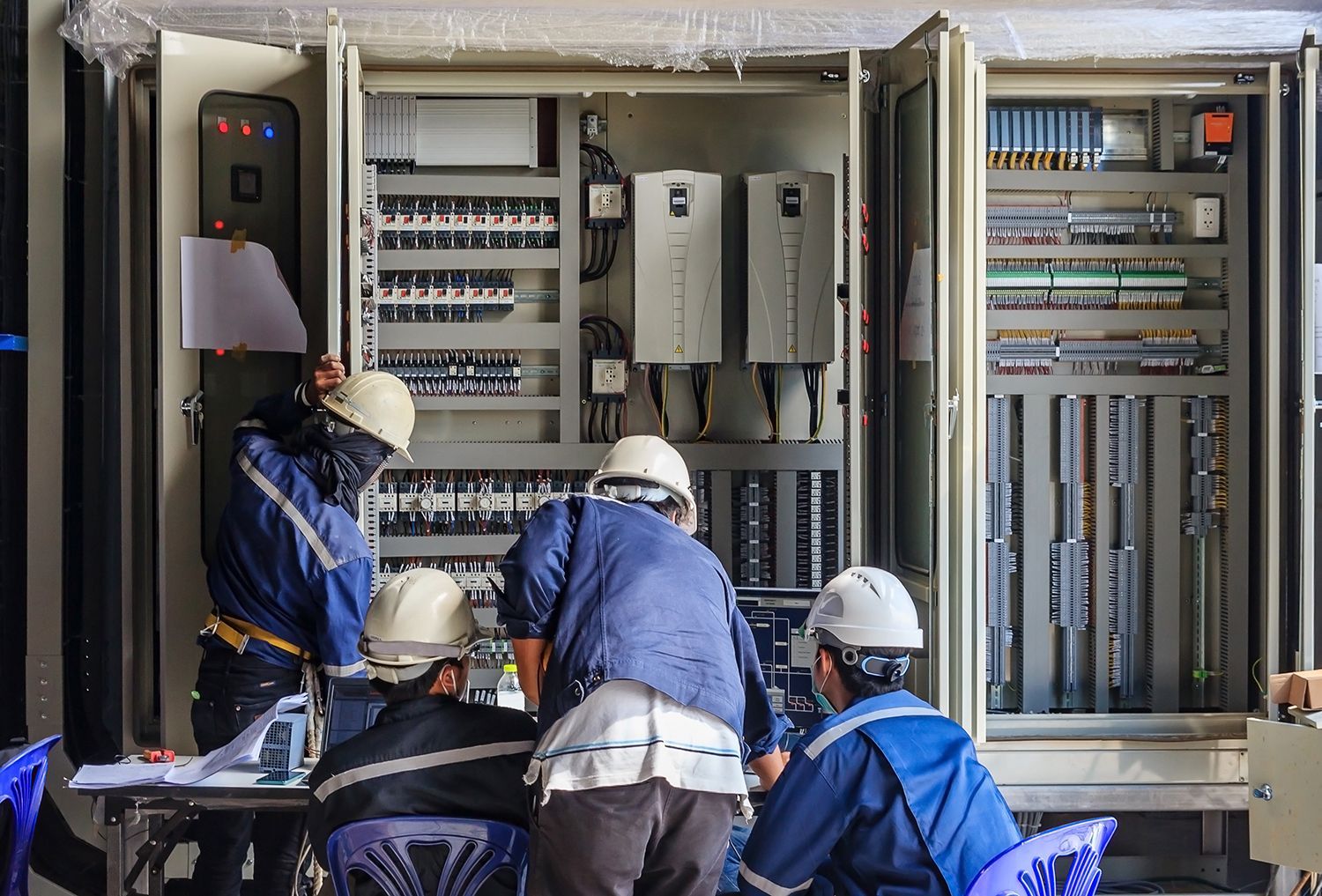 Technicians in blue work on an open electrical control panel with wiring and circuit breakers