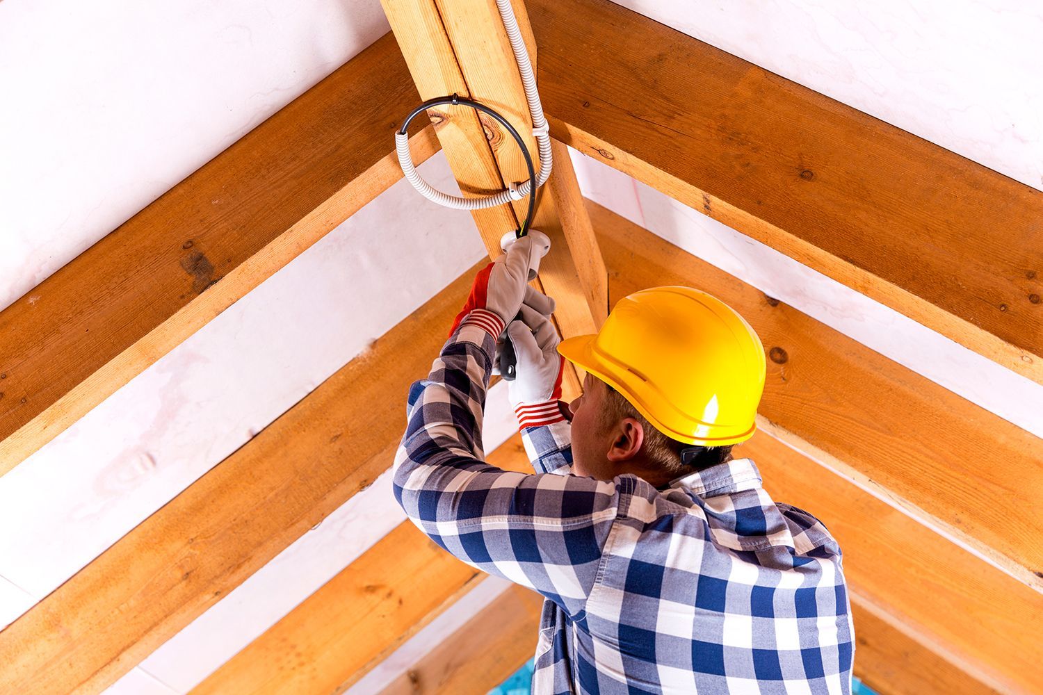 Worker in yellow hard hat installing a ceiling fixture in a wooden room corner.