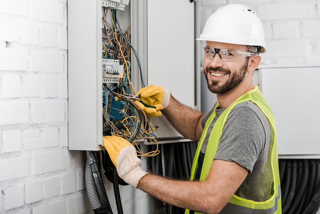 Electrician in hard hat wiring an open electrical panel, wearing gloves and a safety vest.