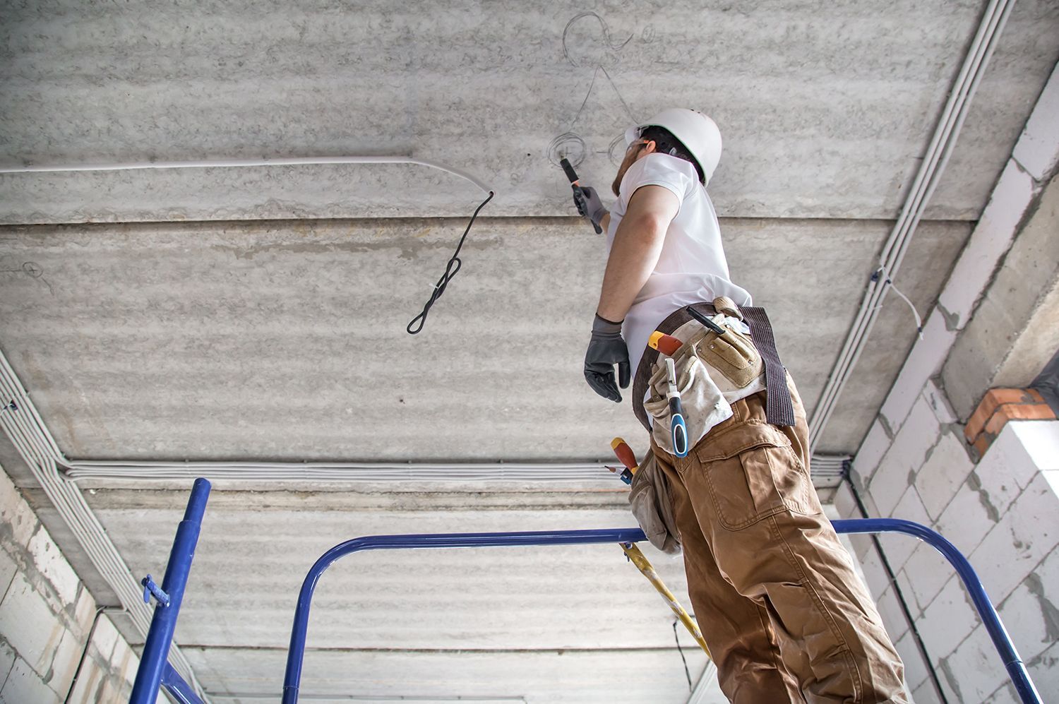 Worker on a ladder painting a ceiling, with blue scaffolding and exposed wiring in a construction area