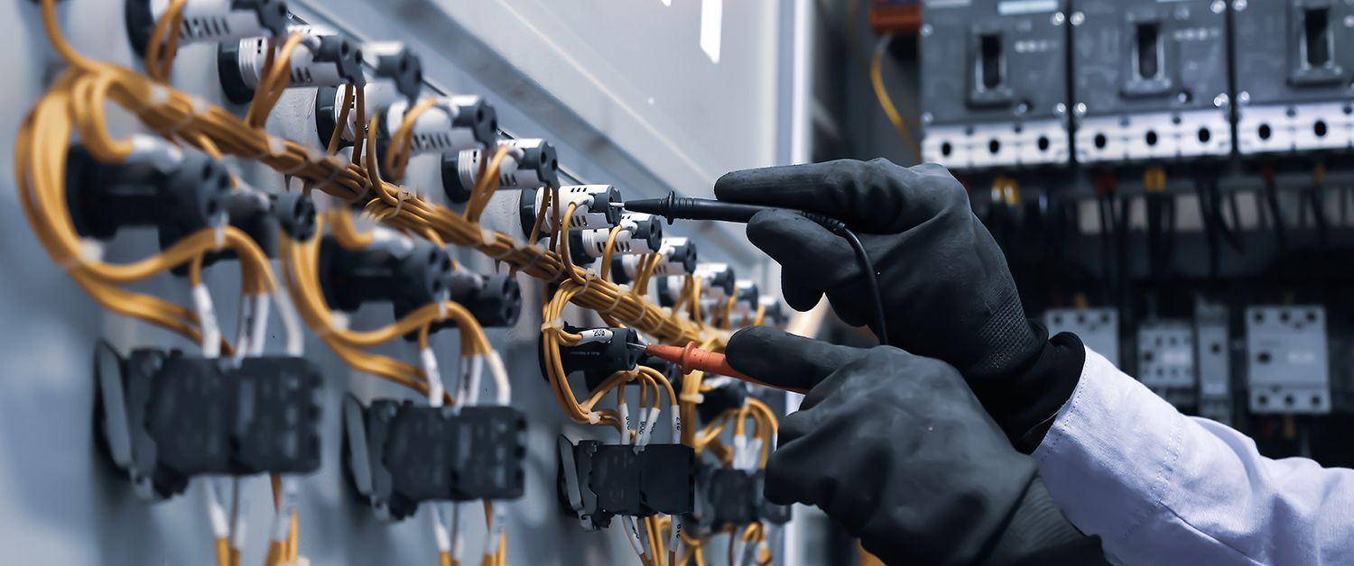 Gloved technician inspects wiring on an electrical control panel with tools in a factory setting