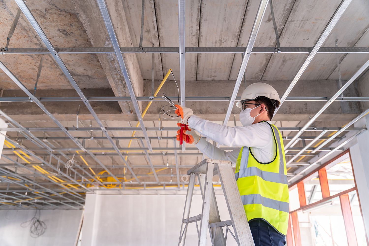 Worker in a hard hat and safety vest installing ceiling fixtures on a ladder in a construction site