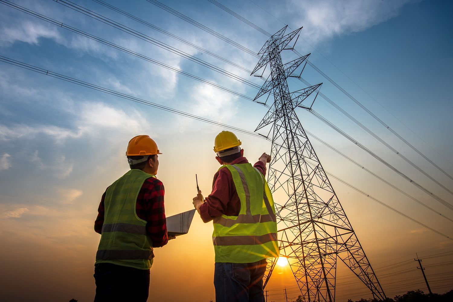 Two utility workers in hard hats inspect a power line tower at sunset.