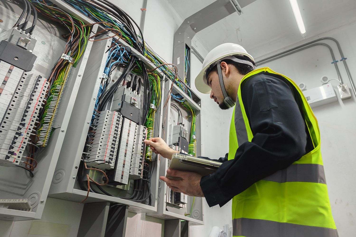 Technician in a hard hat and safety vest inspecting wiring inside an electrical control panel room
